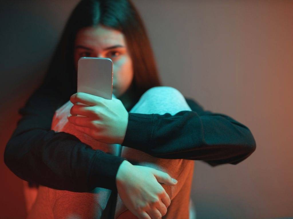 Teenage female sitting and staring at her phone in a darkened environment representing the impact of excessive social media use on mental health.