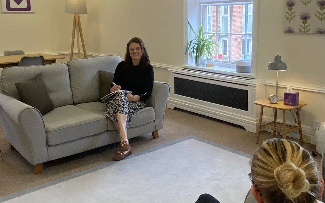 Dr. Danielle Grey of Purple House Clinic sits on a grey sofa in a modern therapy room, smiling.