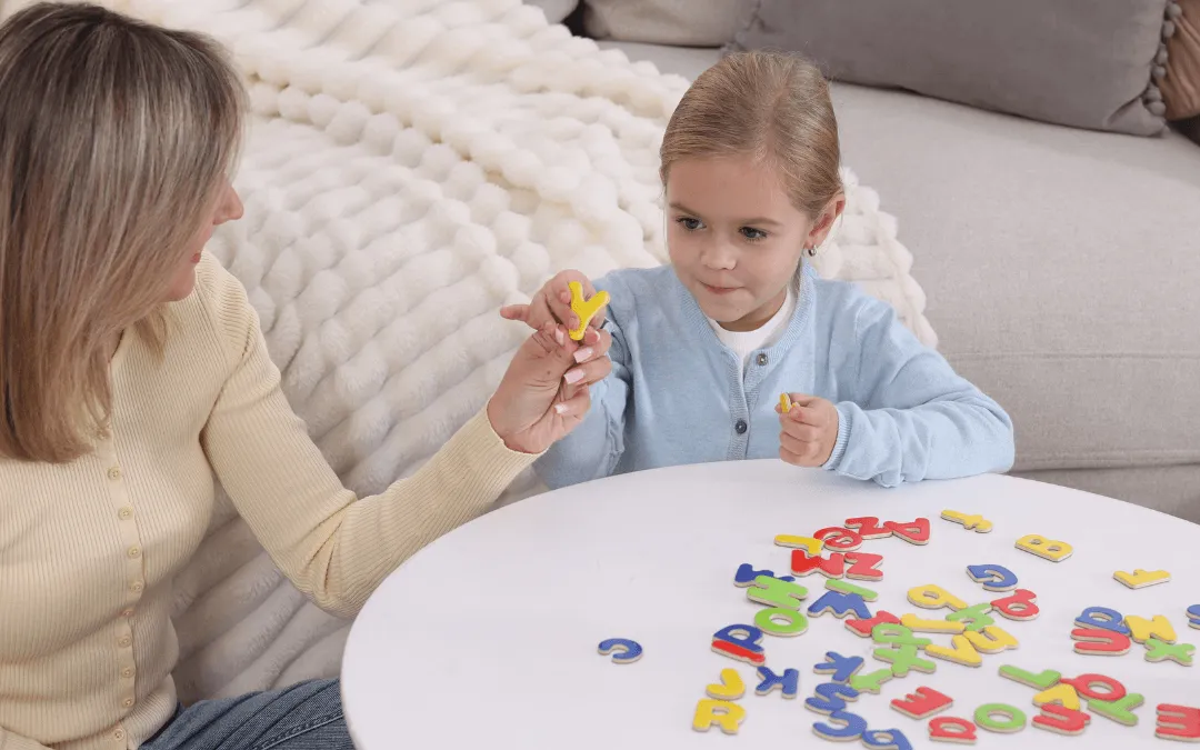 A woman and a young girl sit together on a rug, playing with colourful magnetic letters on a low white circular table.